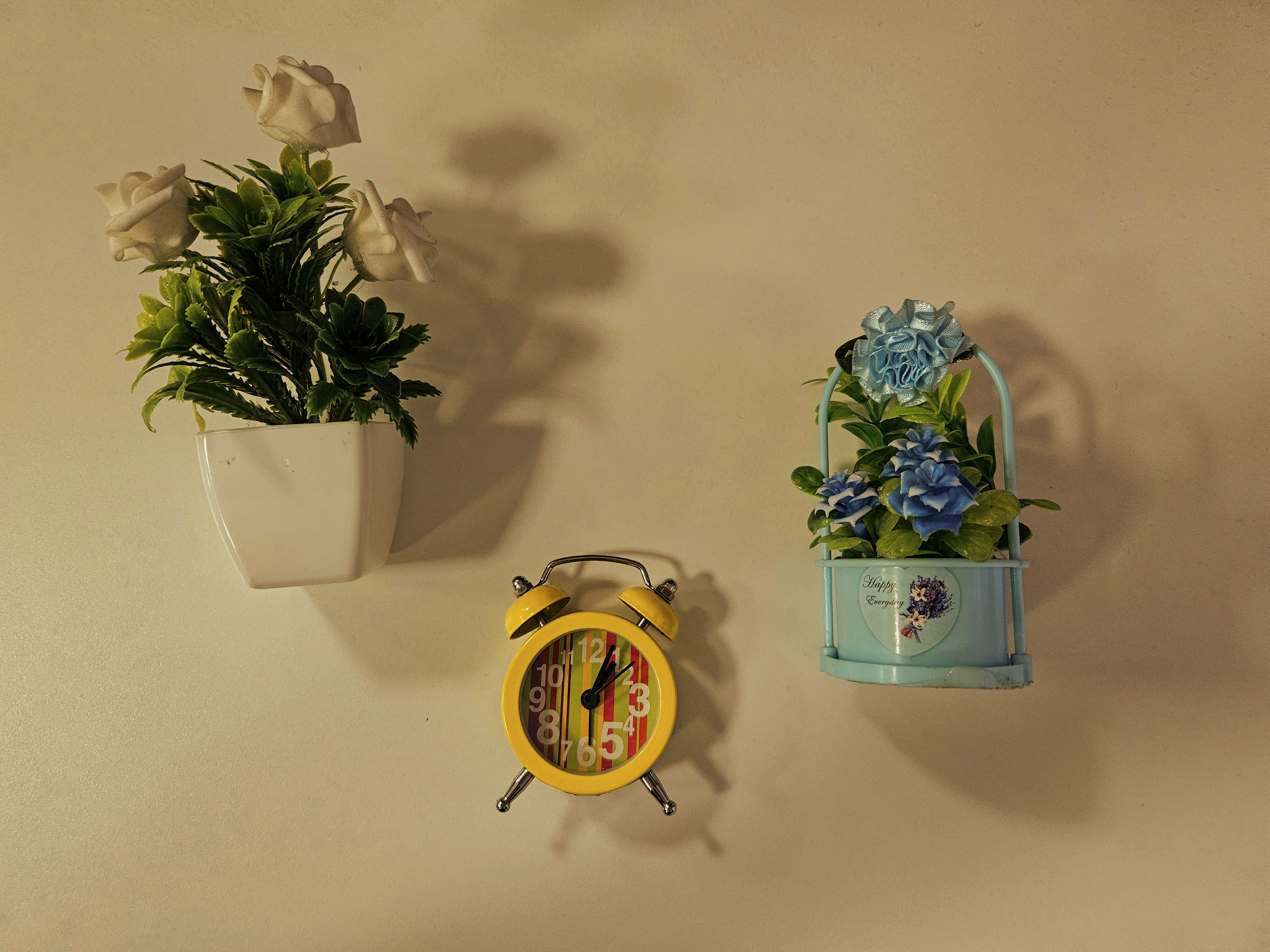 Small wall-mounted planters with flowers and a colorful alarm clock on a warm cream wall