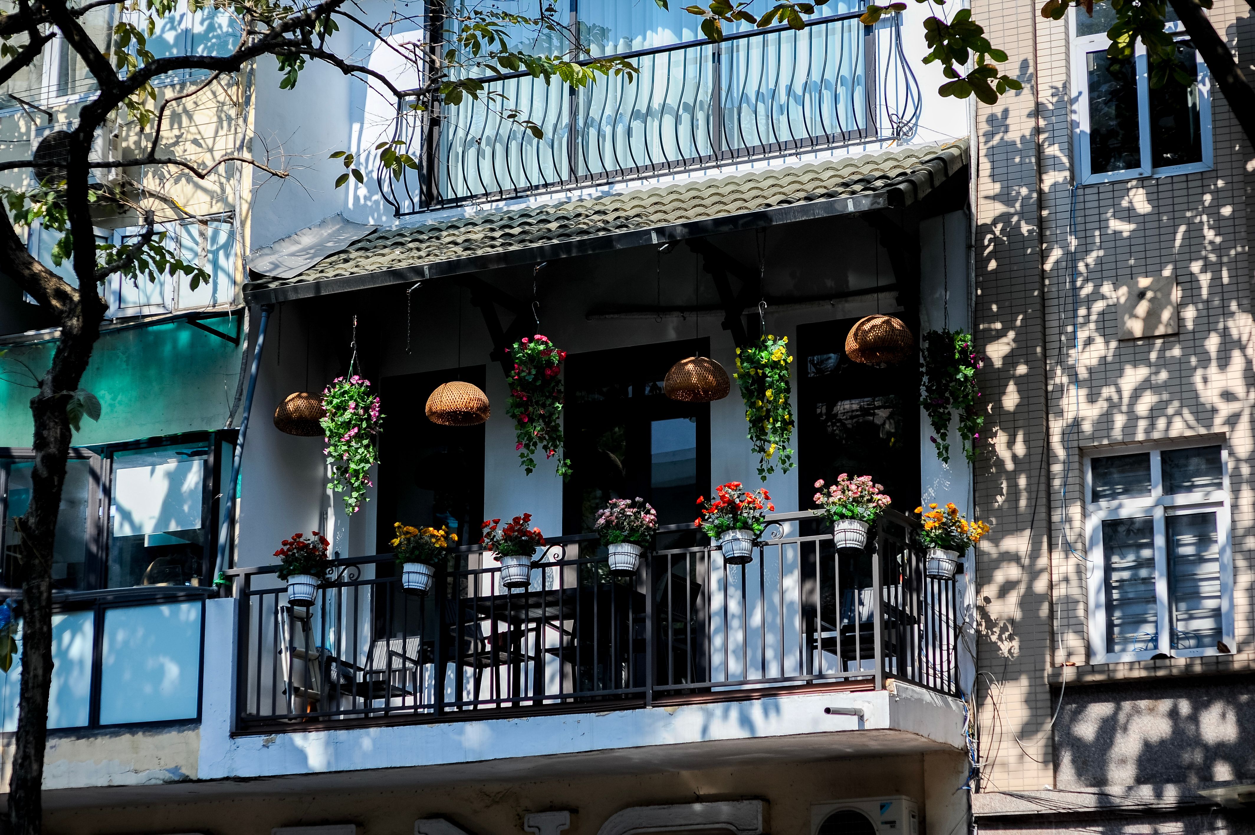 A lush urban balcony overflowing with hanging flower baskets, potted plants, and warm woven pendant lights