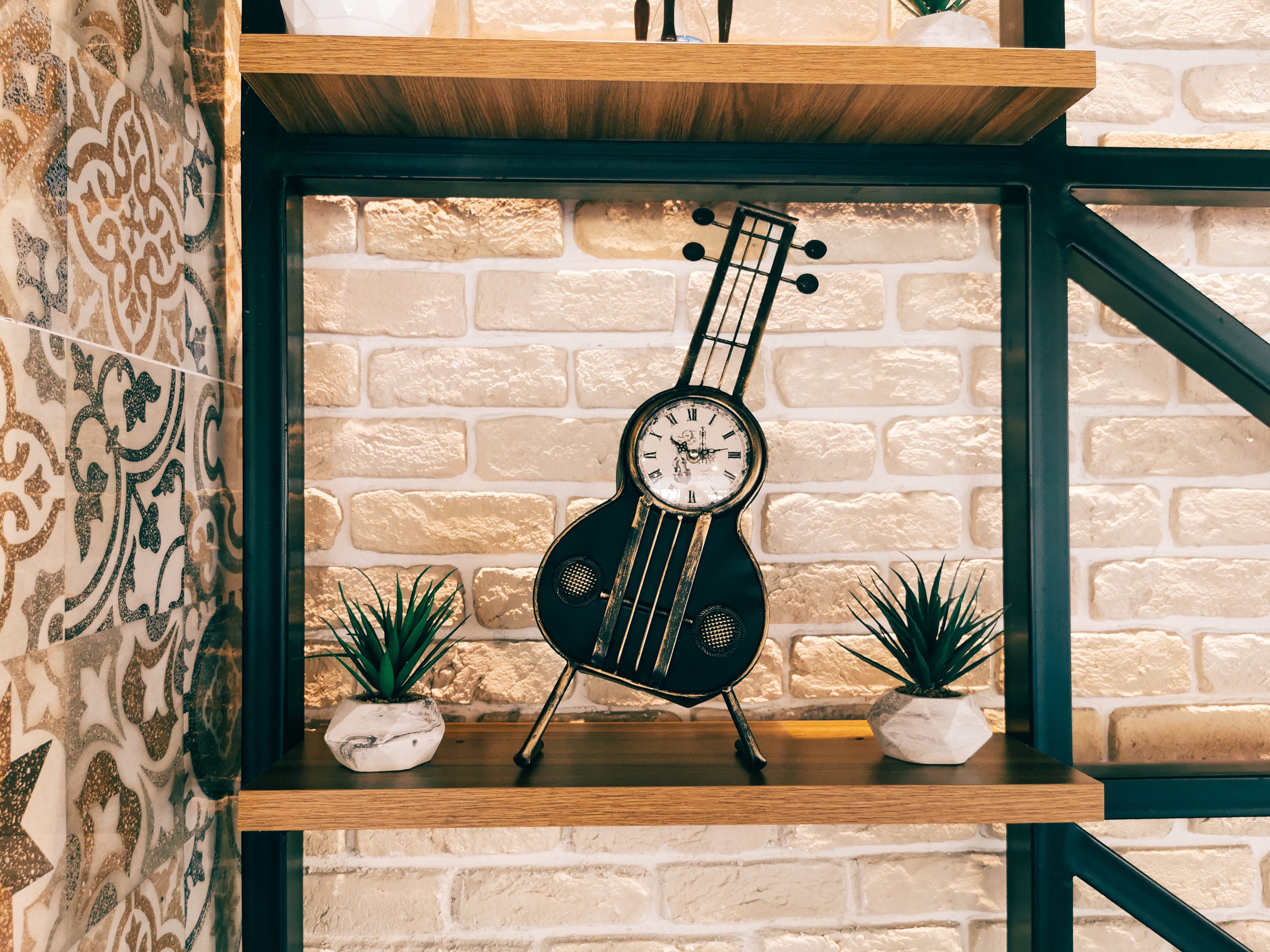 A quirky guitar-shaped vintage clock styled on a wooden shelf with small succulents against a brick wall
